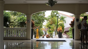 L’entrée d’un hôtel, avec un sol carrelé réfléchissant, une balustrade blanche et une vue sur une végétation luxuriante à l’extérieur.
