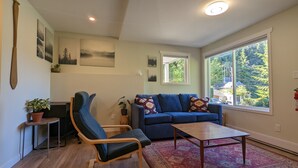 A living room with a blue sofa, a wooden rocking chair, a coffee table, and a large window with a view of trees. - Comfortable one bedroom by the ocean (Cowichan Bay)