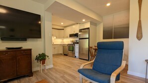 A modern living room with a flat-screen TV, a blue armchair, wooden flooring, and a kitchen in the background. - Comfortable one bedroom by the ocean (Cowichan Bay)