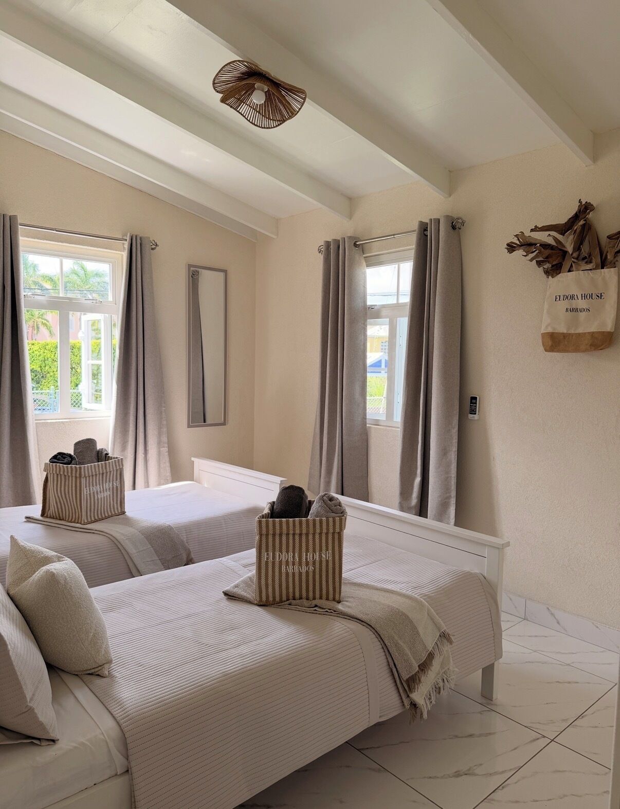 Habitación de hotel con dos camas, un ventanal con cortinas, una lámpara de techo y una cesta de almacenamiento en la pared con la etiqueta 'Eldora House Barrados'.