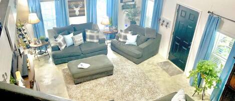 A living room with a sectional sofa, a coffee table, and a small side table. There are blue curtains, a chandelier, and a plant.
