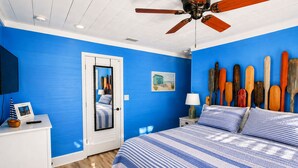 A bedroom with a blue wall, a bed with striped bedding, a ceiling fan, a wooden headboard, and a wall-mounted paddleboard collection. - Clown Fish Cottage - Pet Friendly with King Bed (Mexico Beach)