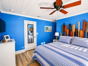 Un dormitorio con pared azul, cama con ropa de cama a rayas, ventilador de techo, cabecera de madera y una colección de tablas de remo fijada a la pared.