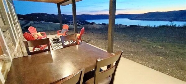 A covered outdoor seating area with a table and chairs, overlooking a lake and hills.