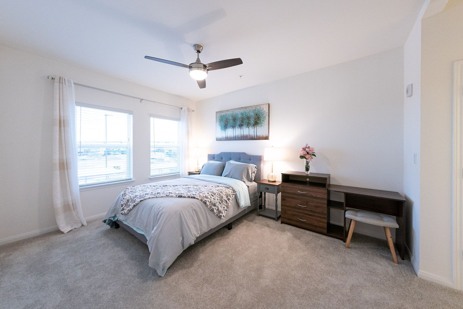 Un dormitorio con cama, mesitas de noche, escritorio, silla, ventilador de techo y ventana con cortinas.