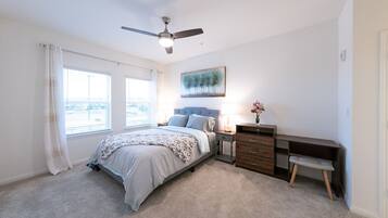 Un dormitorio con cama, mesitas de noche, escritorio, silla, ventilador de techo y ventana con cortinas.