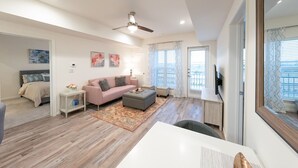 Uma sala de estar moderna com sofá, banquinho e TV em um móvel. Há um ventilador de teto, uma janela com cortinas e uma porta que dá para outro cômodo.