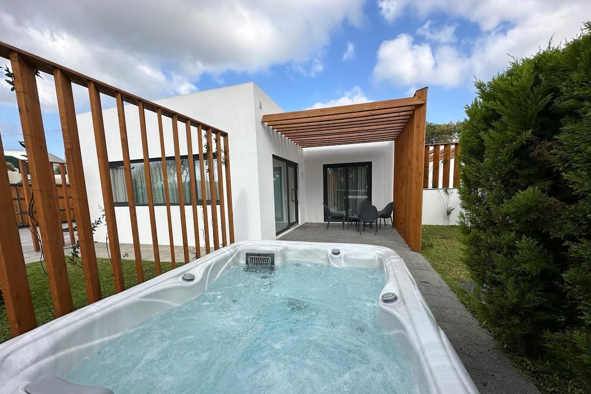 Un jacuzzi en un patio trasero con un cerco de madera, un porche con sillas y una casa moderna al fondo.