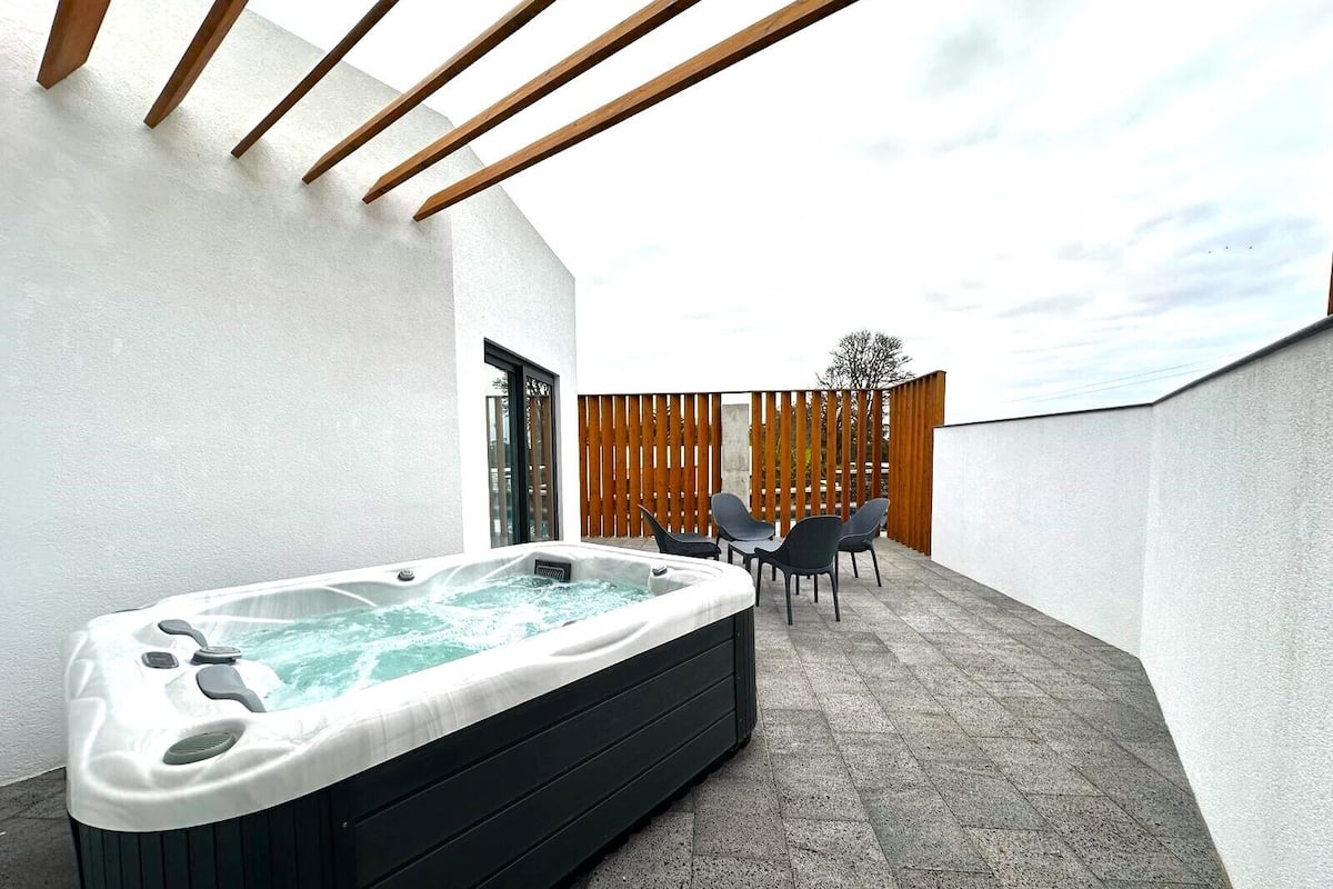 Un jacuzzi en un área exterior cubierta con una pérgola de madera, un espacio de descanso con sillas y una valla de madera al fondo.