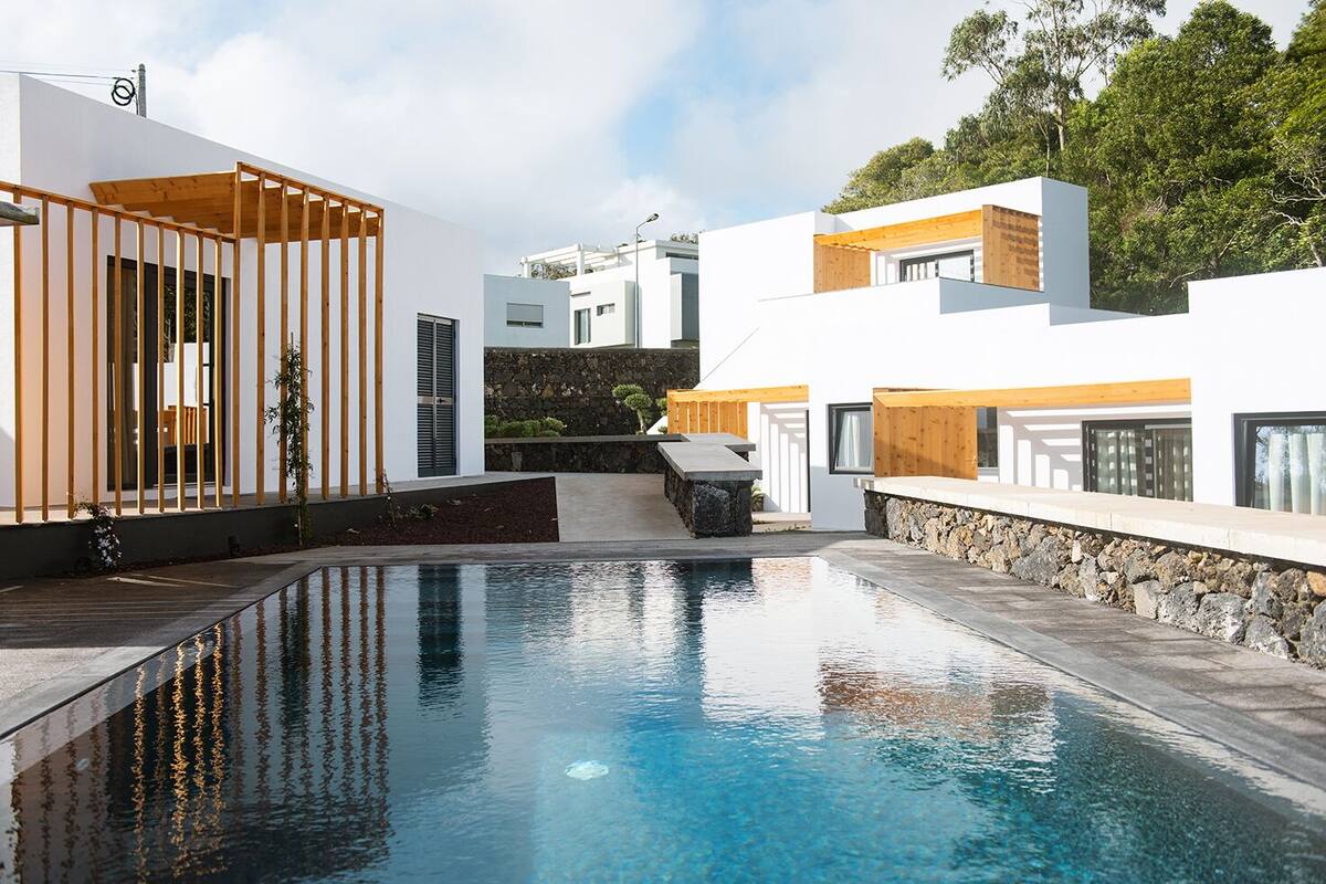 Un área de piscina moderna con una piscina cristalina, pérgolas de madera y edificios blancos con detalles de piedra.