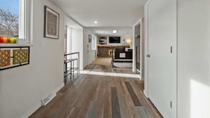 A hallway with wooden flooring, white walls, and a kitchen area visible in the distance.