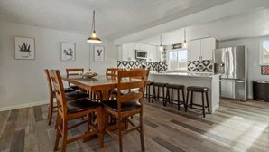 A modern kitchen with a dining area, featuring a wooden table and chairs, a kitchen island with stools, and a stainless steel refrigerator.
