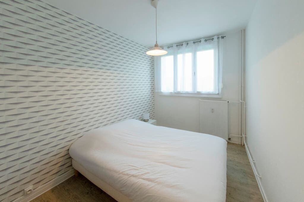A bedroom with a white bed, a textured wall, a window with curtains, and a hanging light fixture.