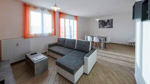 A modern living room with a grey sectional sofa, a small coffee table, and a dining area with a white table and chairs. - Appart'Hôtel L'ile en Mouvement (Montbéliard)