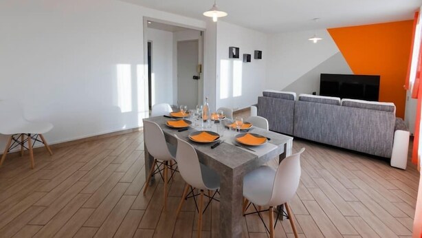 A modern dining area with a long table set for four, surrounded by white chairs with wooden legs. A grey sofa and a television are visible in the background. - Appart'Hôtel L'ile en Mouvement (Montbéliard)
