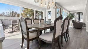 Uma sala de jantar com mesa e cadeiras de madeira, um candelabro e janelas amplas que dão para o exterior.