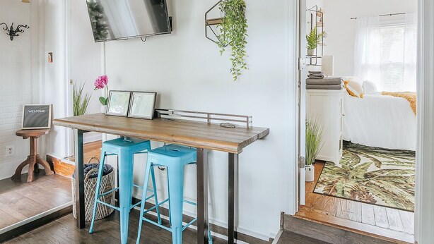 Een moderne woonkamer met een houten eettafel, twee blauwe barstoelen, een wandtelevisie en een hangplant.
