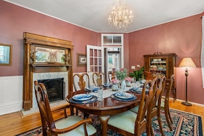 A dining room with a wooden table set for six, surrounded by chairs. A fireplace with a mantelpiece is visible on the left, and a chandelier hangs from the ceiling.