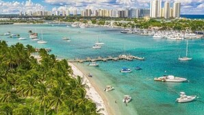 Un paysage urbain côtier avec un port de plaisance, des eaux turquoise et limpides, et une plage bordée de palmiers.