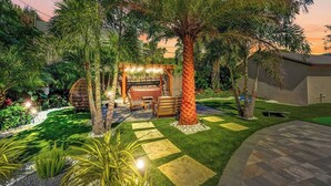Un jardin bien entretenu, agrémenté d’un espace détente, de palmiers et d’une pergola illuminée de guirlandes lumineuses.