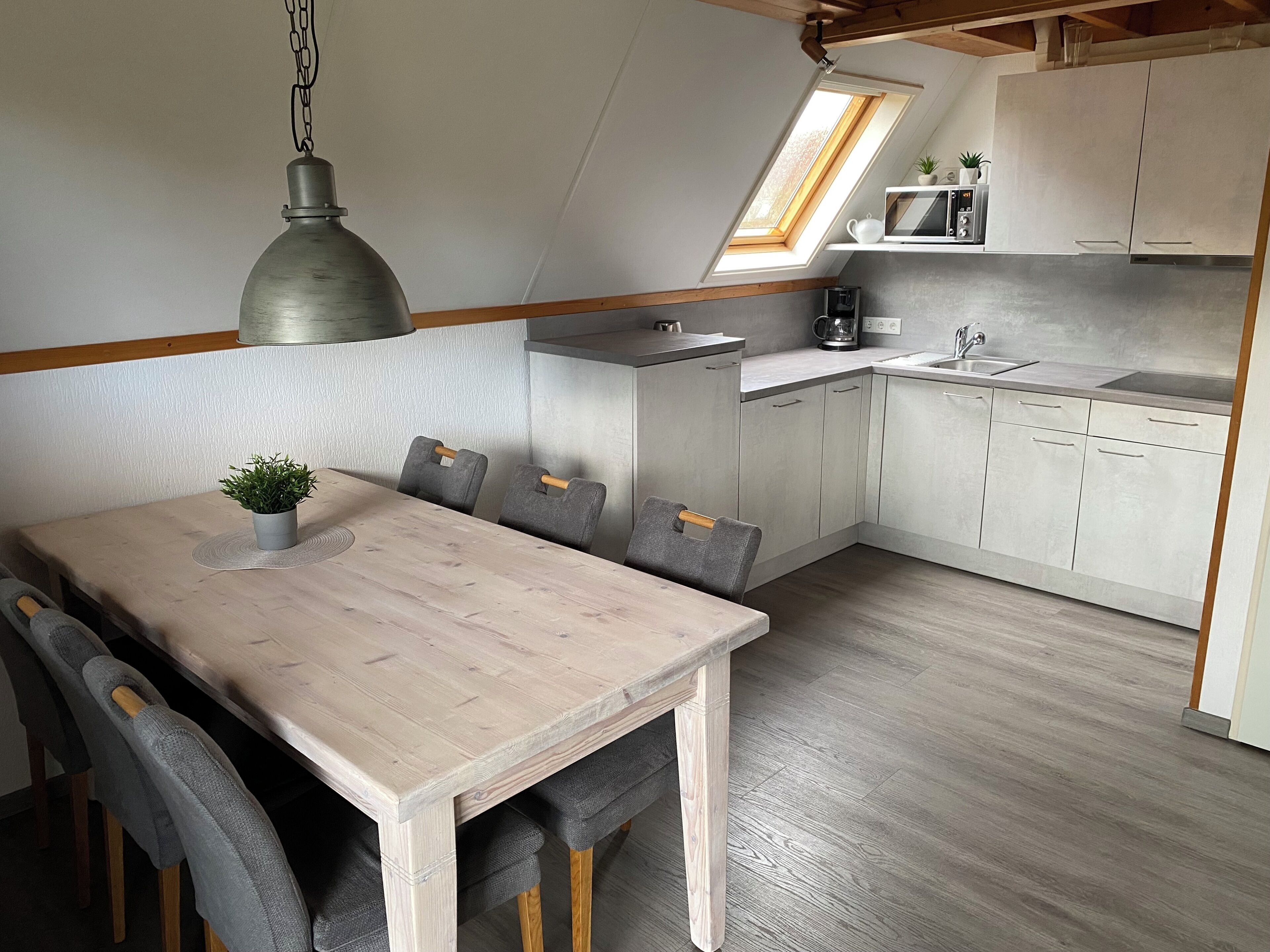 A compact kitchen with a dining area, featuring a wooden table, grey upholstered chairs, a hanging pendant light, and a small potted plant.