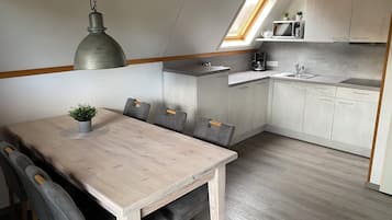 A compact kitchen with a dining area, featuring a wooden table, grey upholstered chairs, a hanging pendant light, and a small potted plant.