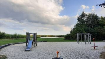 Une aire de jeux avec un toboggan et des balançoires, entourée d’une zone sablonneuse et de verdure.