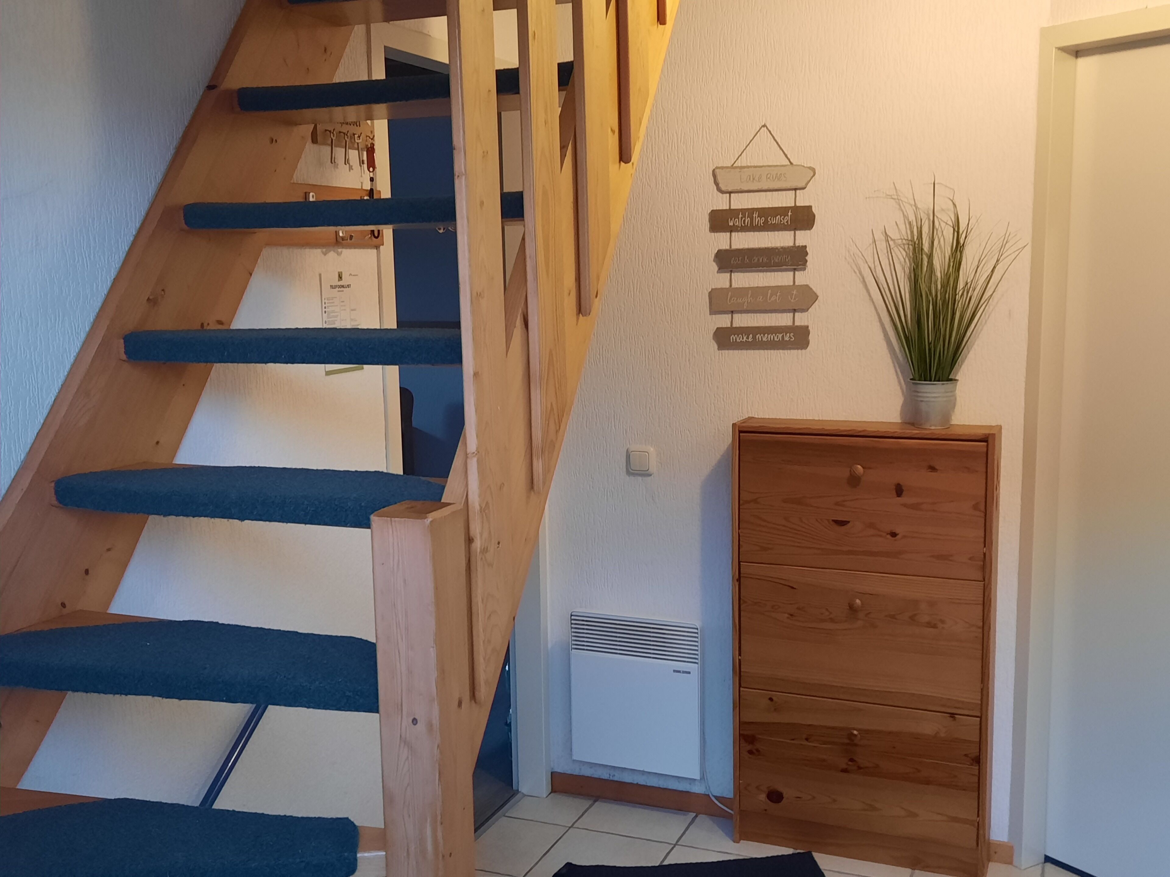 Een trap met blauwe treden, een houten dressoir en een bord aan de muur met zinnen als 'geniet van de zonsondergang' en 'creëer herinneringen'.