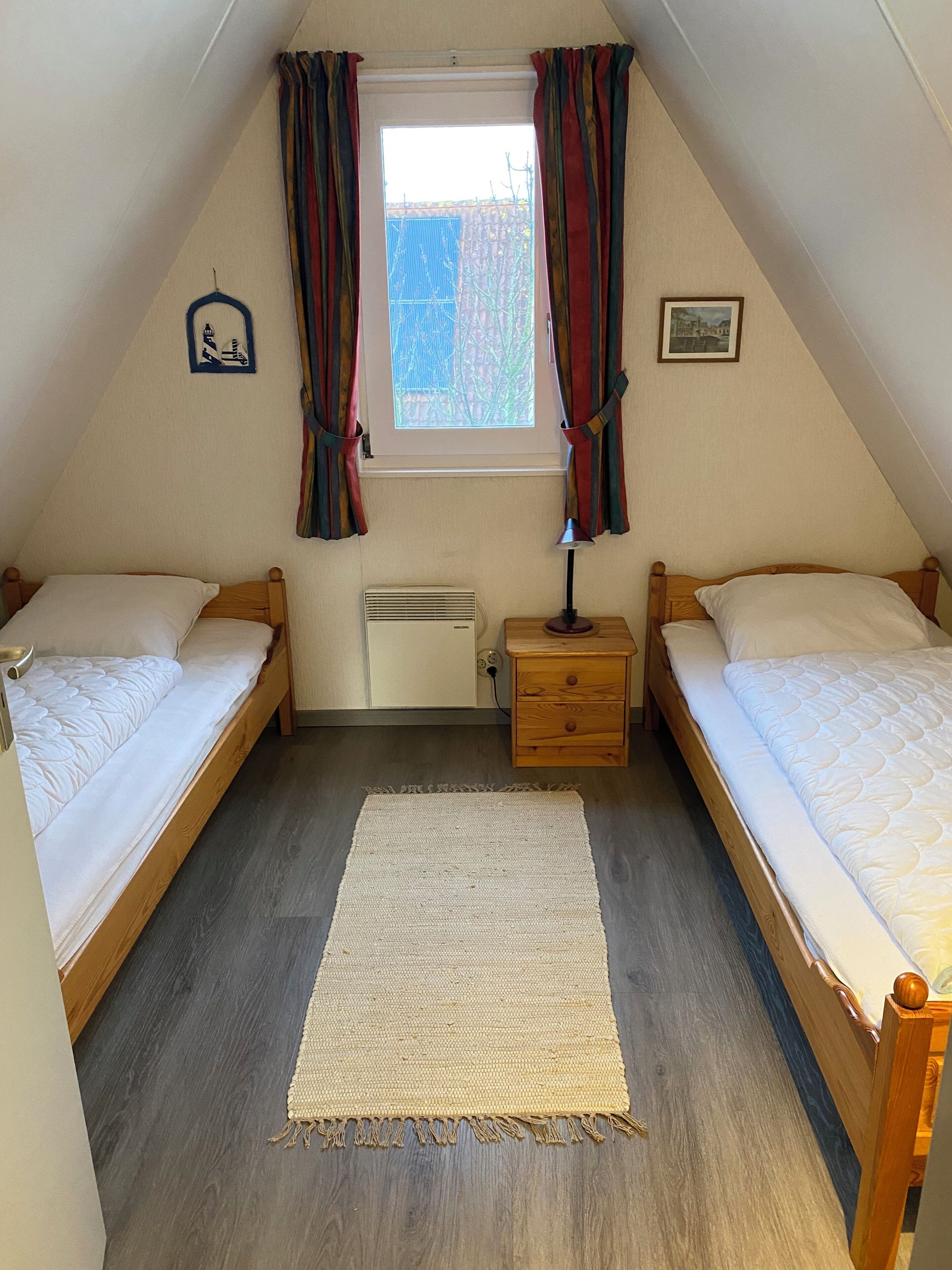 Een knus kamertje met twee houten bedden, een raam met gordijnen, een radiator en een klein nachtkastje met een lamp.