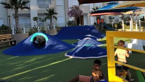 A playground with slides and climbing structures, surrounded by a building with balconies and a palm tree.