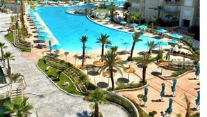 A resort with a large central pool, multiple swimming areas, palm trees, and a modern building complex.