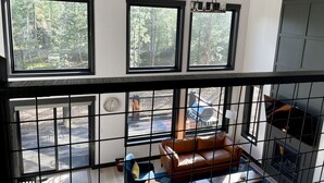 Une pièce avec vue sur une forêt, grâce à de grandes fenêtres, un canapé en cuir marron et une petite table avec une lampe.
