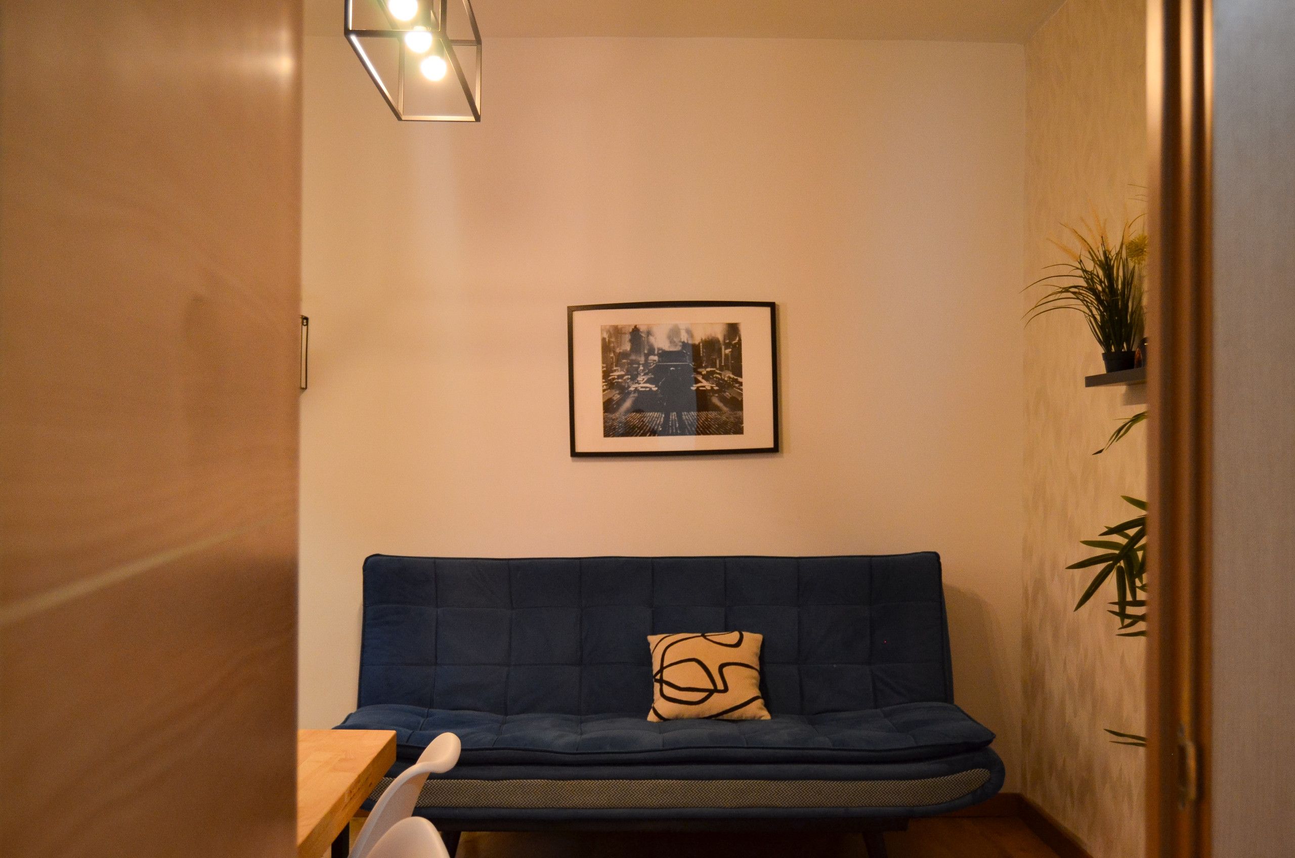 Ett rum med en blå soffa, en inramad tavla på väggen och en krukväxt till höger.