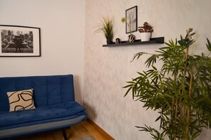 Huoneessa on sininen sohva, seinällä on kehystetty taulu ja hyllyllä on kasveja sekä koriste-esineitä.