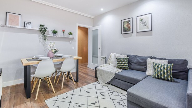 Una sala moderna con un sofá gris, una mesa de comedor con sillas y una pequeña zona de cocina.