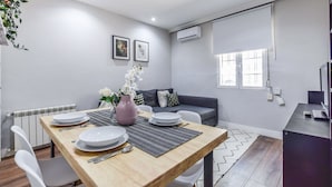 Una sala de estar moderna con una mesa de comedor preparada para cuatro personas, un sofá gris y un amplio ventanal con persianas.