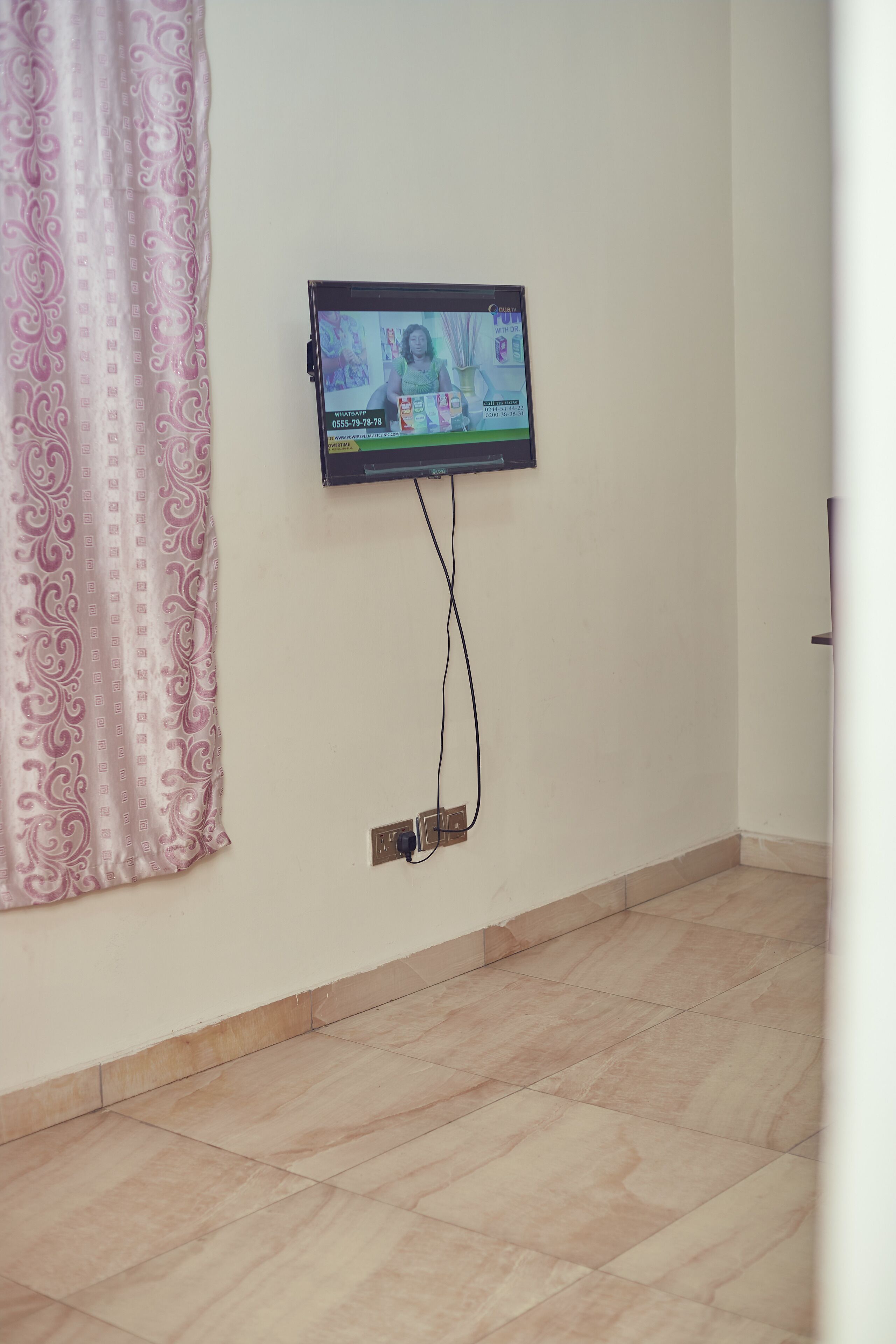 A room with a television mounted on the wall, a patterned curtain, and a tiled floor.