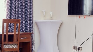Una habitación con una silla de madera, una pequeña mesa blanca, un televisor en la pared y una cortina estampada.