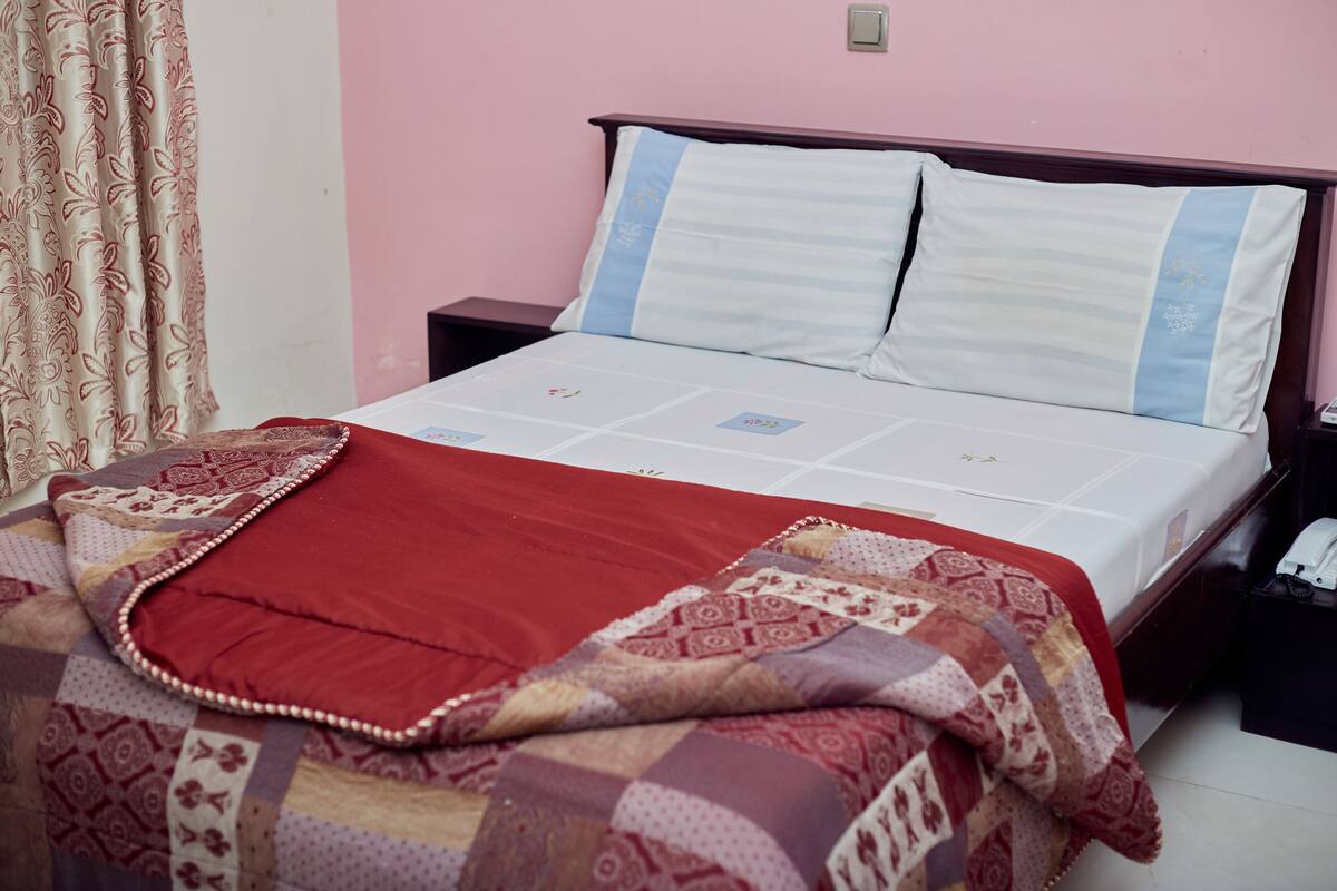 Een keurig opgemaakt bed met een rode deken en een geborduurde quilt, een houten hoofdbord en een nachtkastje met een telefoon.