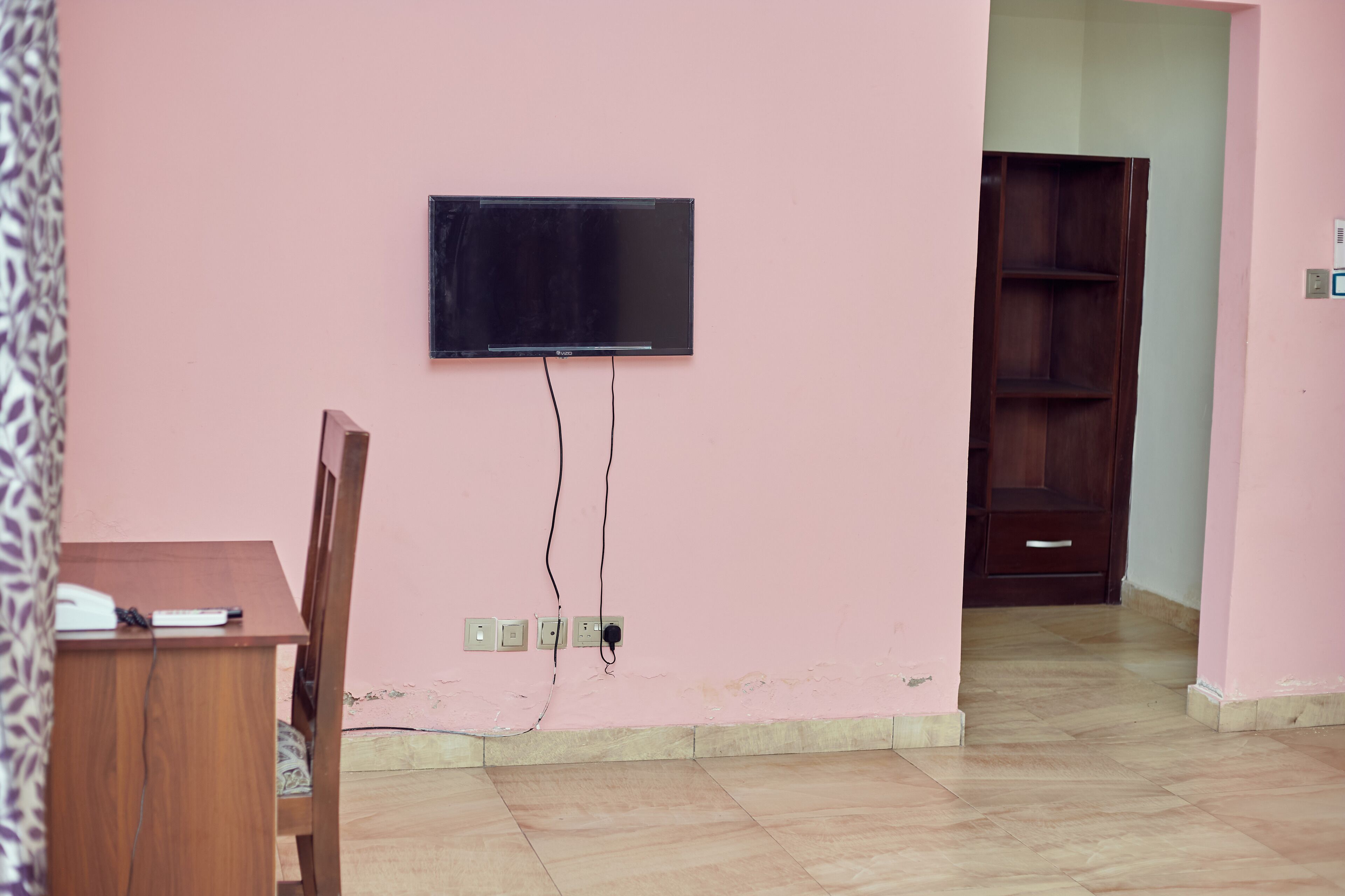 Una habitación con televisión montada en la pared, un escritorio de madera con teléfono y un armario de madera.