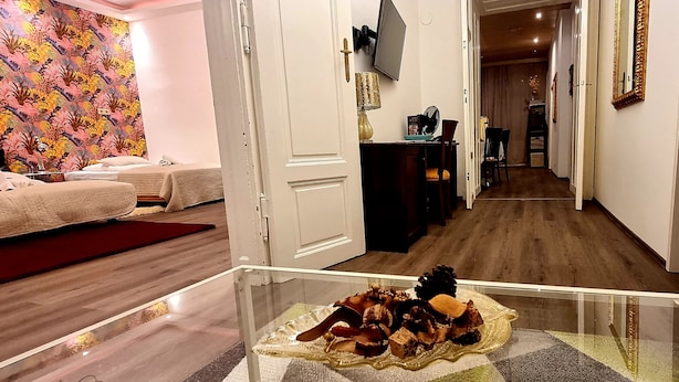 A hotel room with a bed, a glass table with decorative items, a TV, and a hallway leading to a dining area.