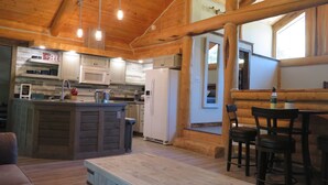Interior acogedor de una cabaña con comedor de madera, cocina con electrodomésticos blancos y un amplio ventanal que proporciona luz natural.