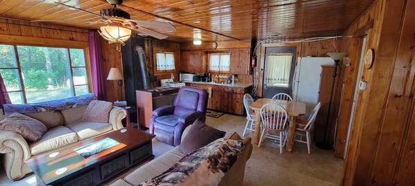 Interior de cabana com sofá, poltrona, mesa de jantar e cozinha.