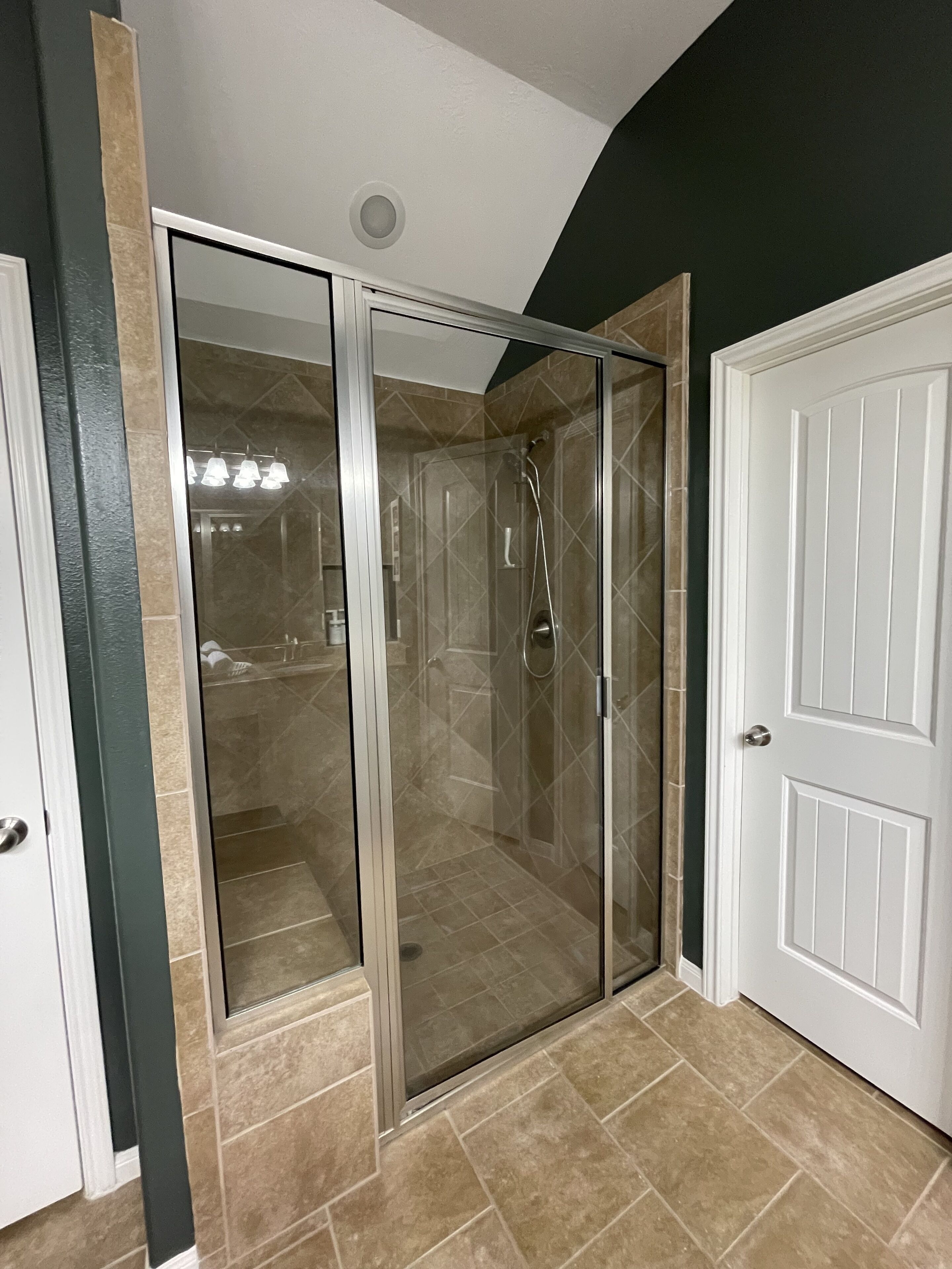 A glass-enclosed shower with a white door, beige tiles, and a white door next to it.