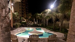Un área de piscina nocturna con varias piscinas, sillas de descanso y palmeras.
