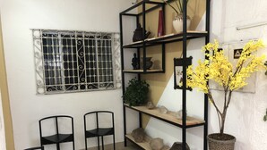 Una habitación con ventana de rejas, dos sillas negras, un estante de madera con adornos y una mesita.
