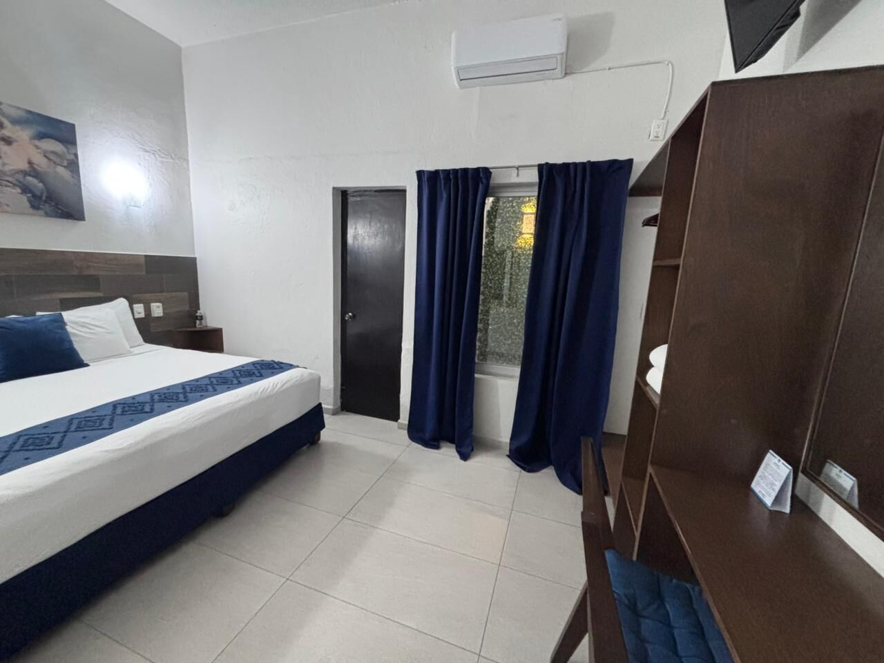 Quarto de hotel com uma cama, janela com cortinas, guarda-roupa de madeira e ar condicionado fixado na parede.