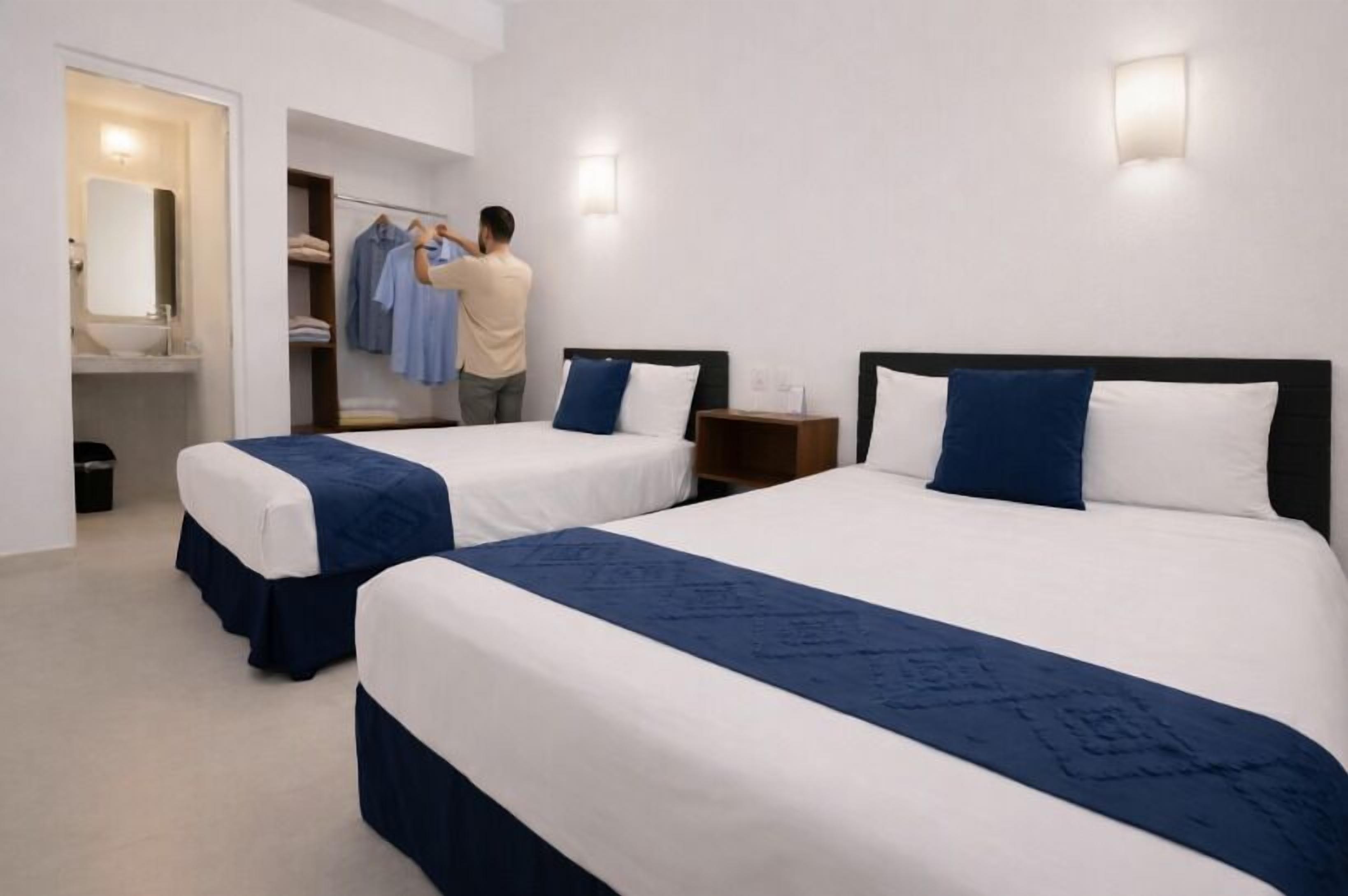 A hotel room with two beds, a bathroom, and a person hanging clothes.