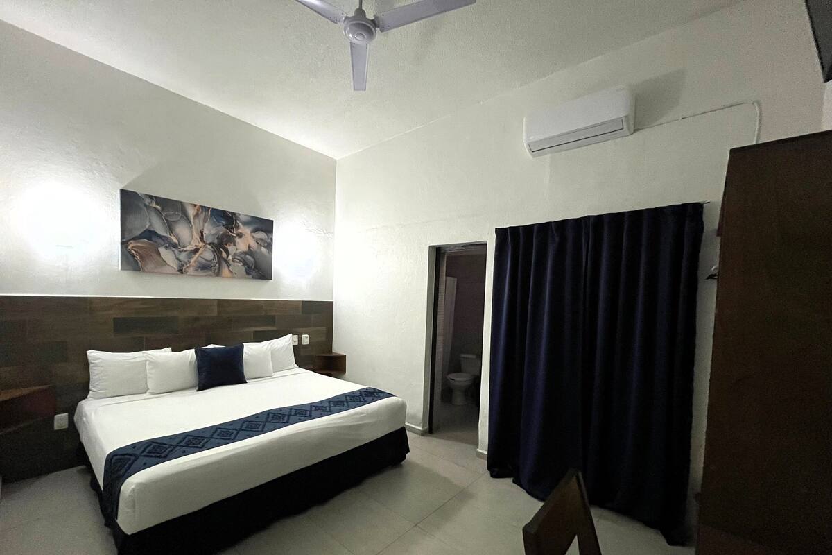 A hotel room with a bed, a ceiling fan, a painting on the wall, and a bathroom visible through an open door.