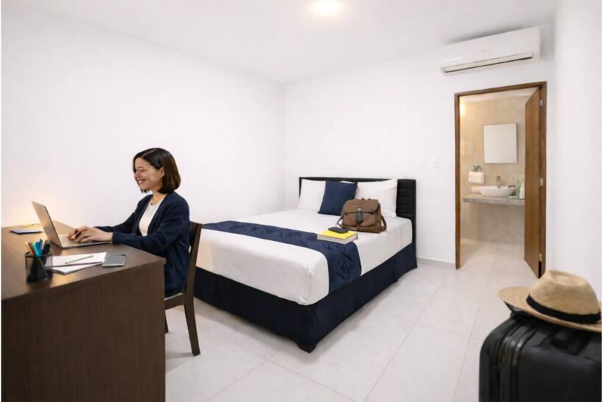 A hotel room with a bed, a desk with a laptop, a suitcase, and a suitcase with a hat.